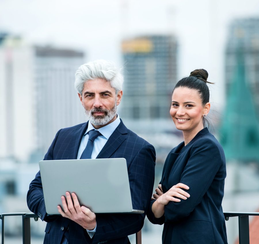 A mature businessman and young businesswoman with laptop standing outdoors on a terrace, working.